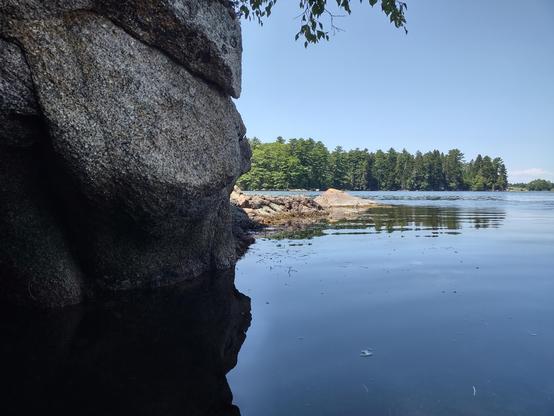 Peeking out from behind a granite outcropping you can see the tidal river flowing by, blue from the reflected sky. The water this side of some partially submerged boulders is calm and smooth, but rippled from wind and the tide beyond. Oak and pine trees line the far shore.