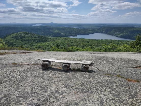 A bench made of a weathered wood plank on three logs sitting on an expanse of smooth rock. It looks down over forests and a series of ponds leading to Penobscot Bay. White puffy clouds dot the blue sky.